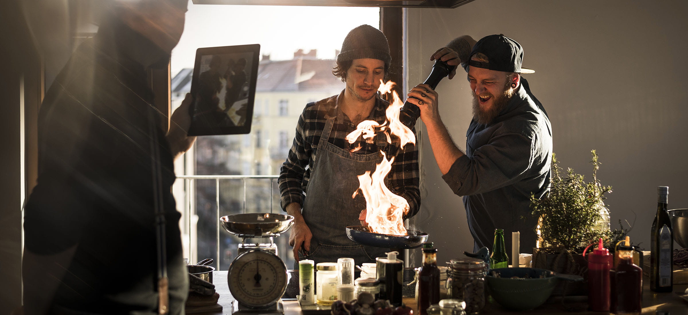 Two people cook in a sunlit kitchen as flames rise from a frying pan, while another person records the scene with a tablet.